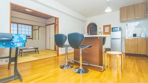 A modern kitchen with wooden cabinets, a refrigerator, a microwave, and a coffee maker. There are two black bar stools and a small round table. - AMP FLAT Nishijin5 (Fukuoka)