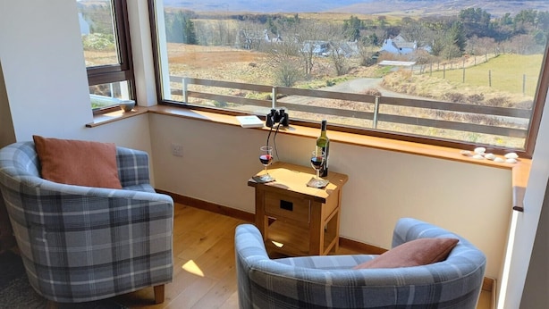 Une pièce avec deux fauteuils à carreaux, une petite table en bois sur laquelle se trouvent un verre et une bouteille de vin, et une vue sur un paysage rural par une grande fenêtre.