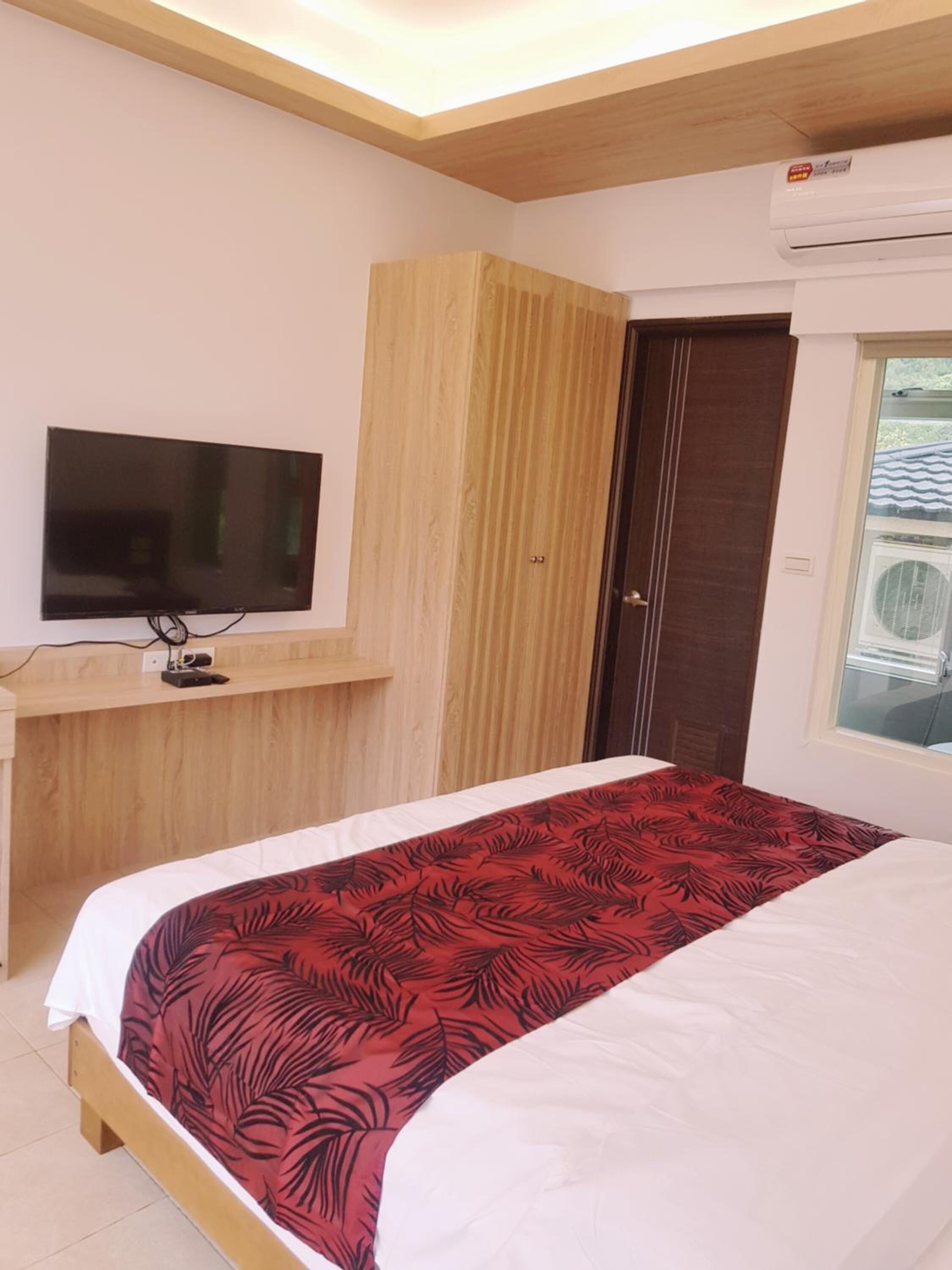 A bedroom with a bed, a TV on a wooden desk, a wardrobe, and an air conditioning unit.