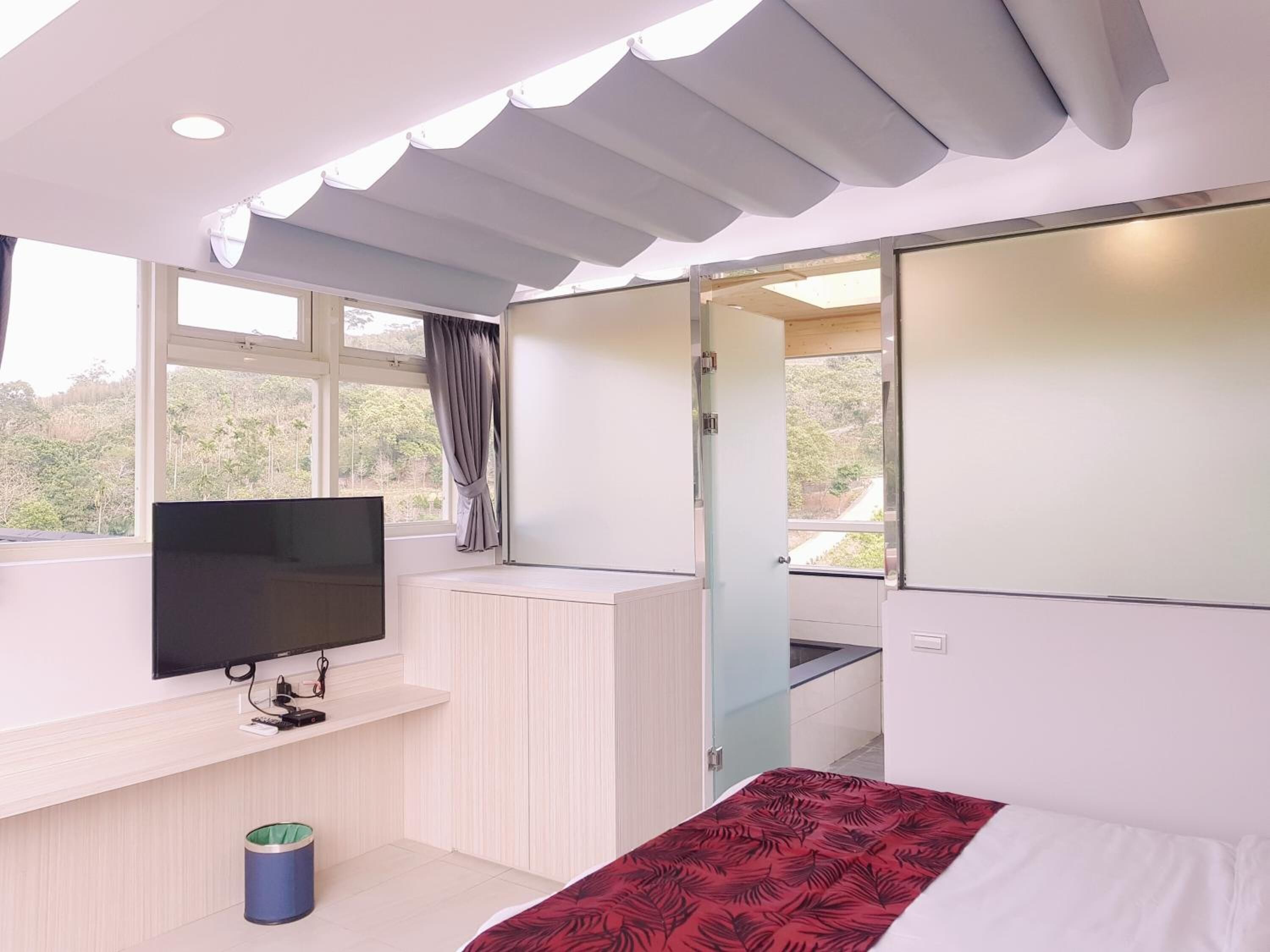 Un dormitorio moderno con una televisión, una cama con colcha estampada roja, un cubo de basura pequeño y una ventana con cortinas.