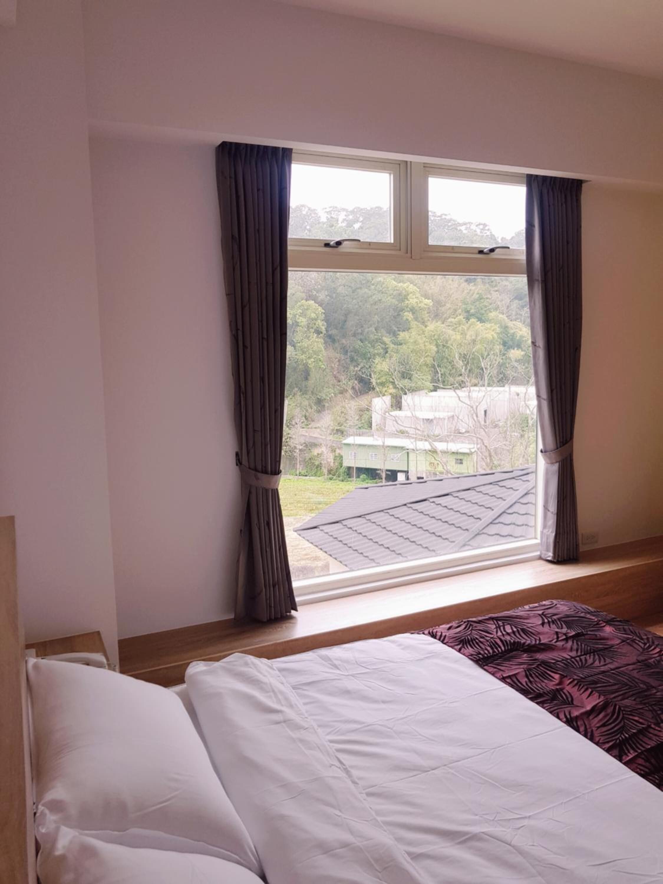 A bedroom with a bed, a window with curtains, and a view of a building and trees outside.