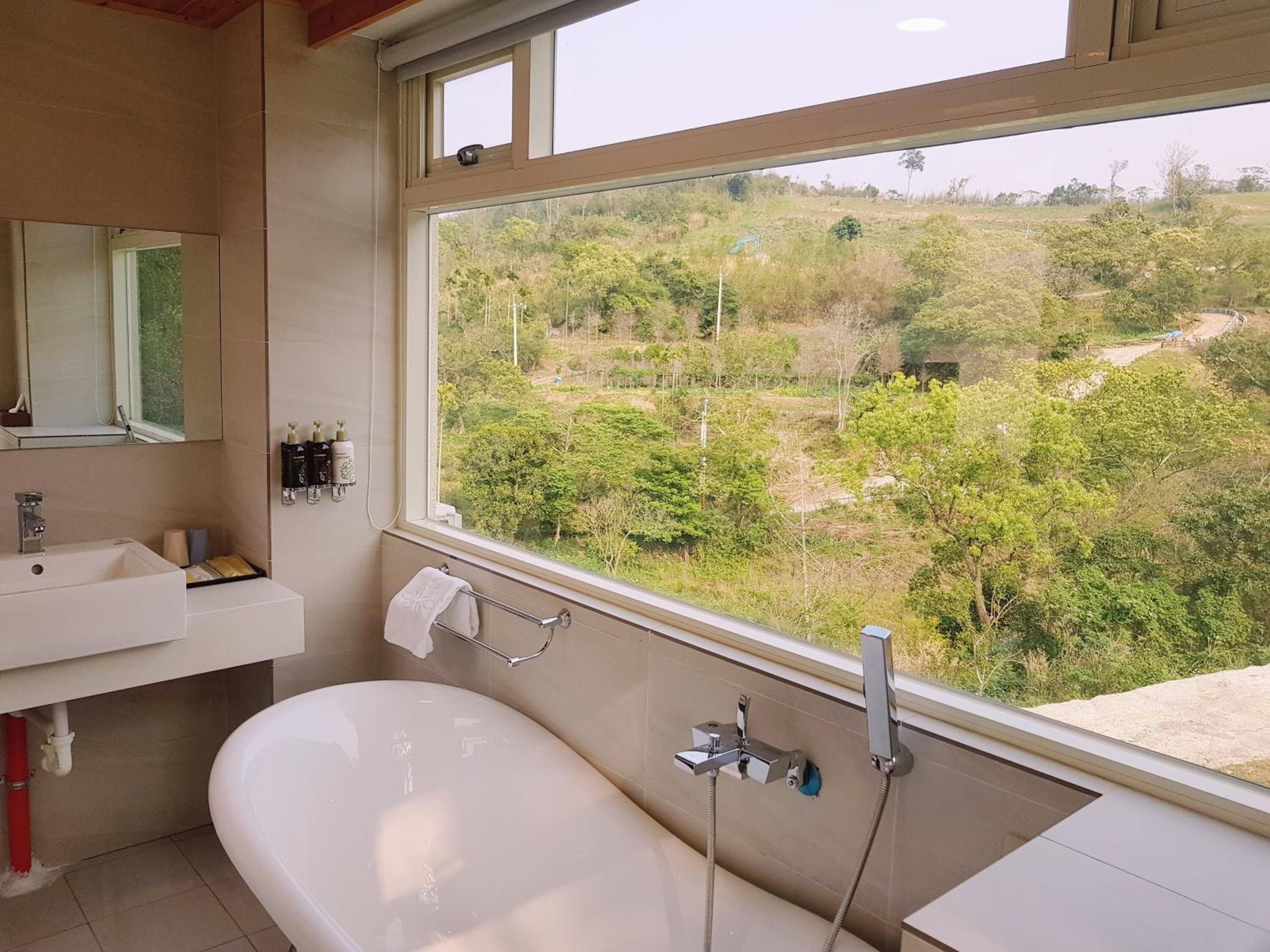 A bathroom with a bathtub, a sink, and a window offering a view of a green landscape.