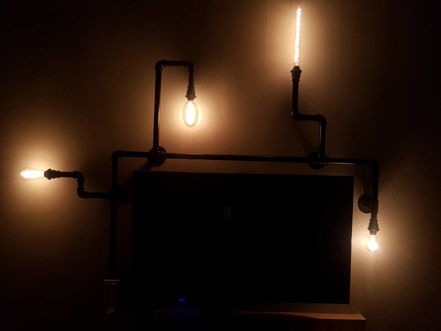 Un televisor con marco negro y pantalla oscura, rodeado por cuatro focos conectados mediante tuberías negras.