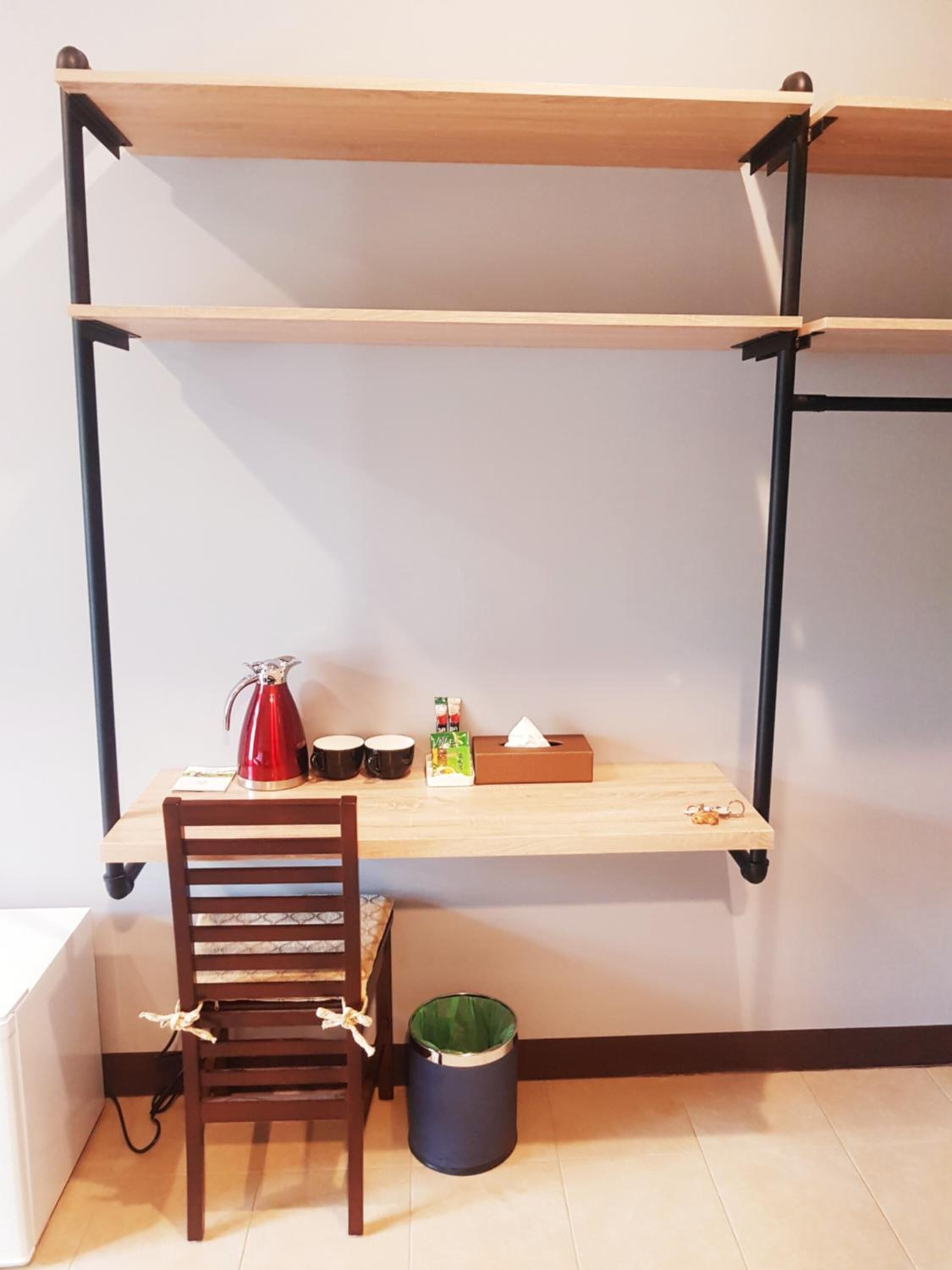 Una habitación pequeña con un escritorio de madera, una silla, una tetera roja, dos tazas negras, una caja de pañuelos y un cubo de basura.