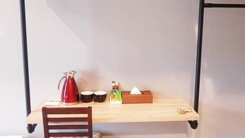 Una habitación pequeña con un escritorio de madera, una silla, una tetera roja, dos tazas negras, una caja de pañuelos y un cubo de basura.