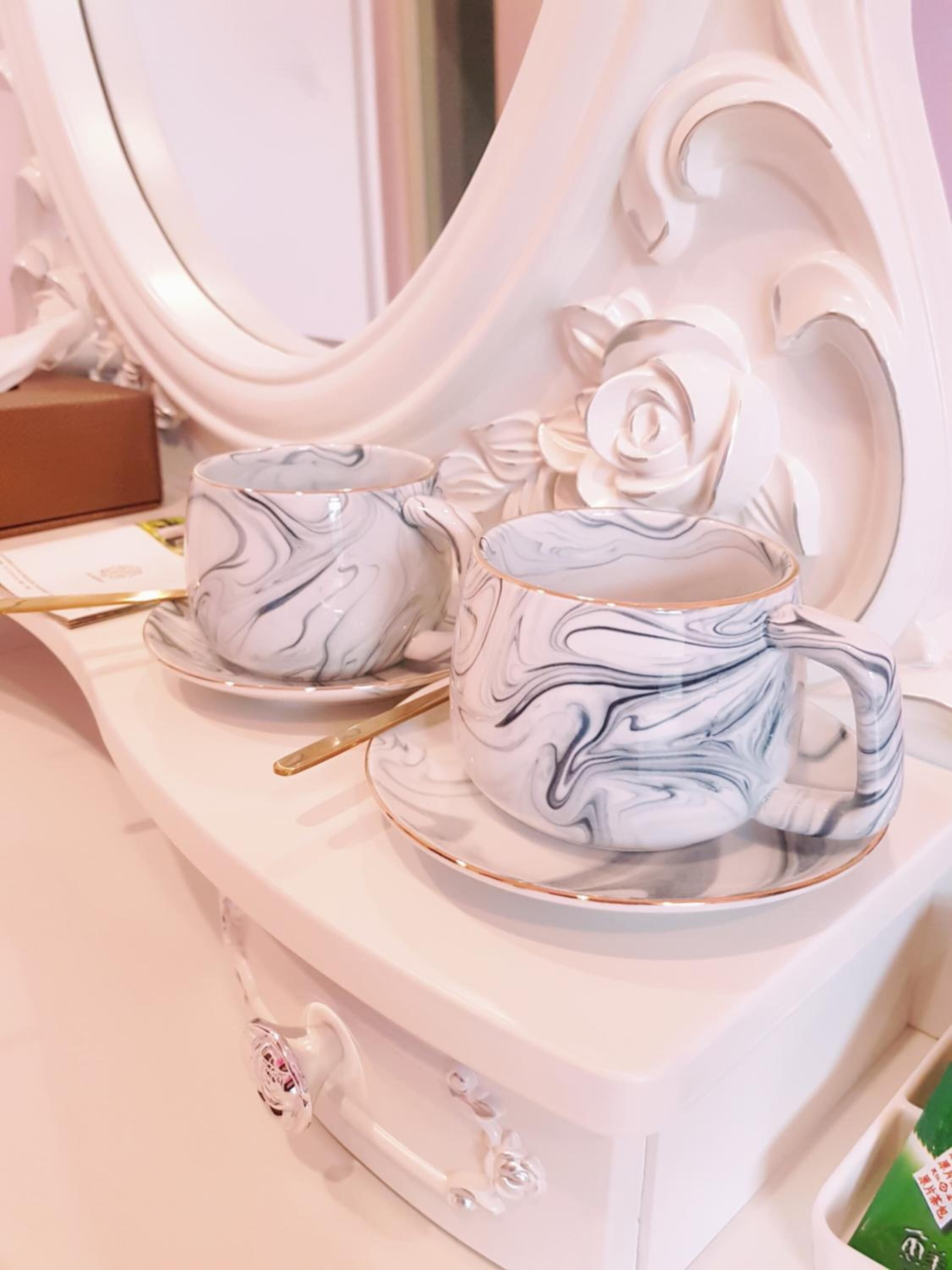 Un tocador blanco con un juego de té de diseño marmolado, que incluye tetera y tazas, y un espejo con marco decorativo.