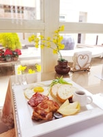 Bandeja de desayuno con huevos, tocino, tostadas y fruta sobre una mesa, cerca de una ventana que da a vistas de edificios y una planta al exterior.