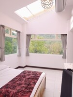 Un dormitorio con una cama grande, dos ventanas con cortinas, un tragaluz y vistas al exterior con vegetación.