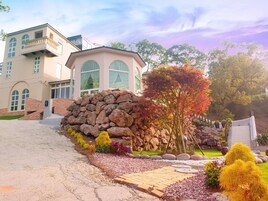 Una casa grande de dos pisos con un muro de piedra y un jardín con diversas plantas y un sendero pavimentado.
