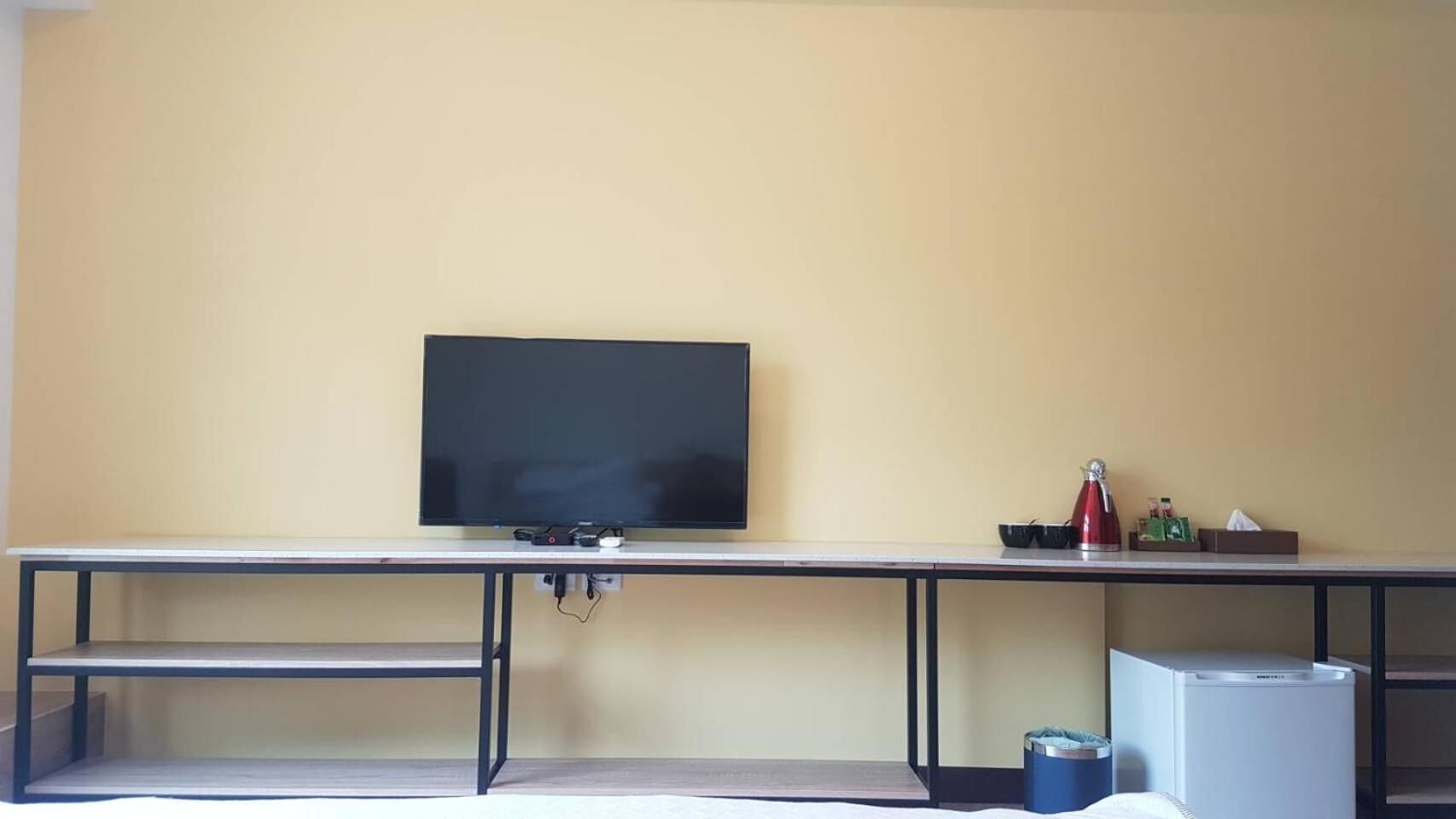 A room with a flat-screen TV mounted on a wall, a desk with a computer, and a small trash bin.