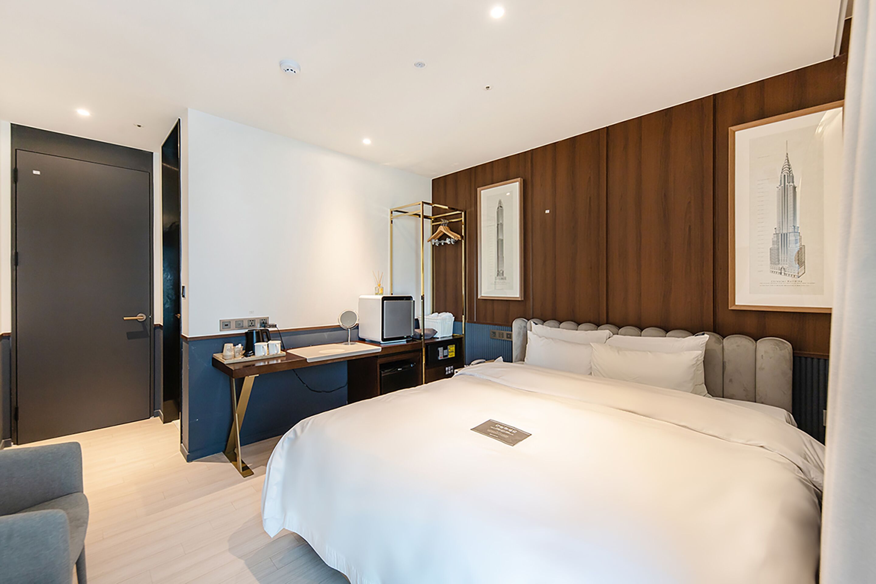 A modern hotel room with a large bed, a desk with a computer, a mirror, and a framed picture on the wall.