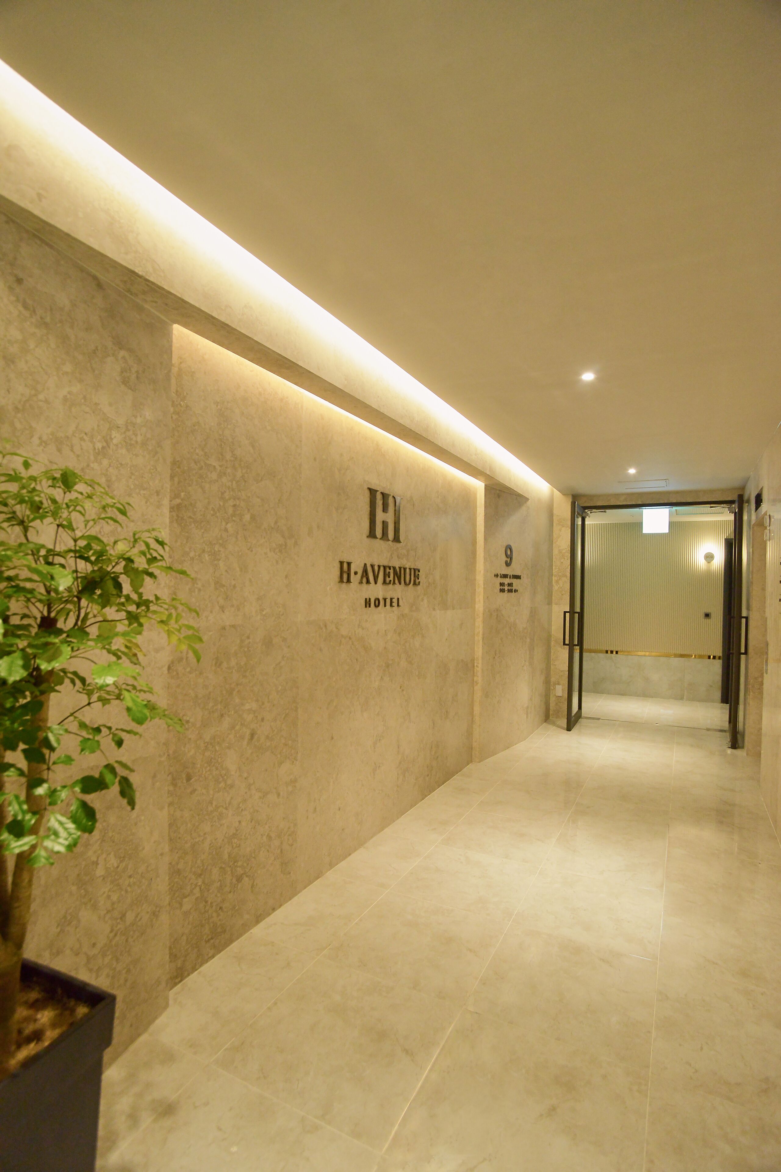 Un pasillo con suelo y paredes de mármol que conduce a una puerta con un cartel que dice 'H-Avenue Hotel'.