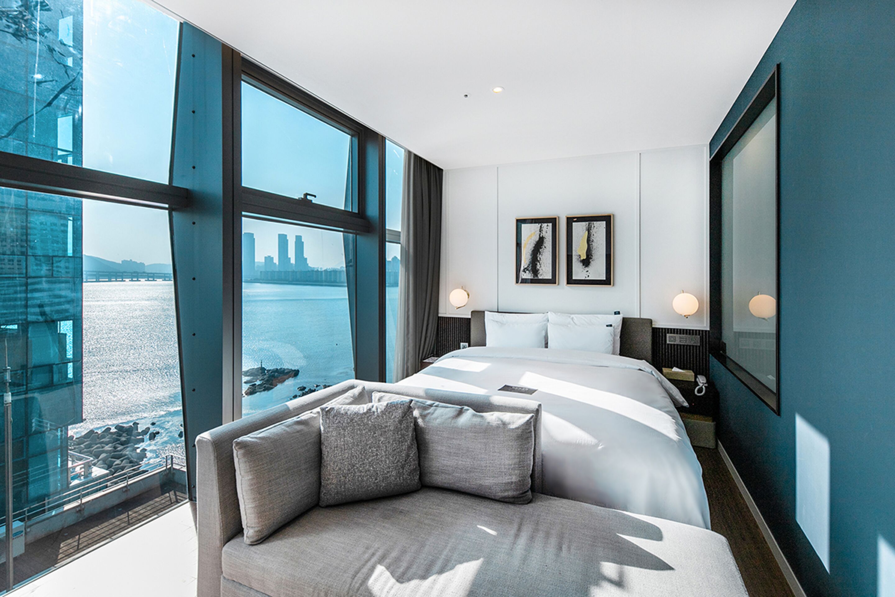 A modern hotel room with a large bed, a sofa, and a view of a cityscape through large windows.