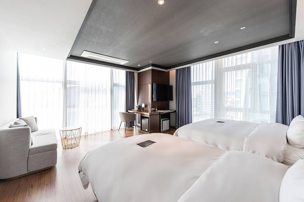 A modern hotel room with two large beds, a desk, a TV, and a sofa.