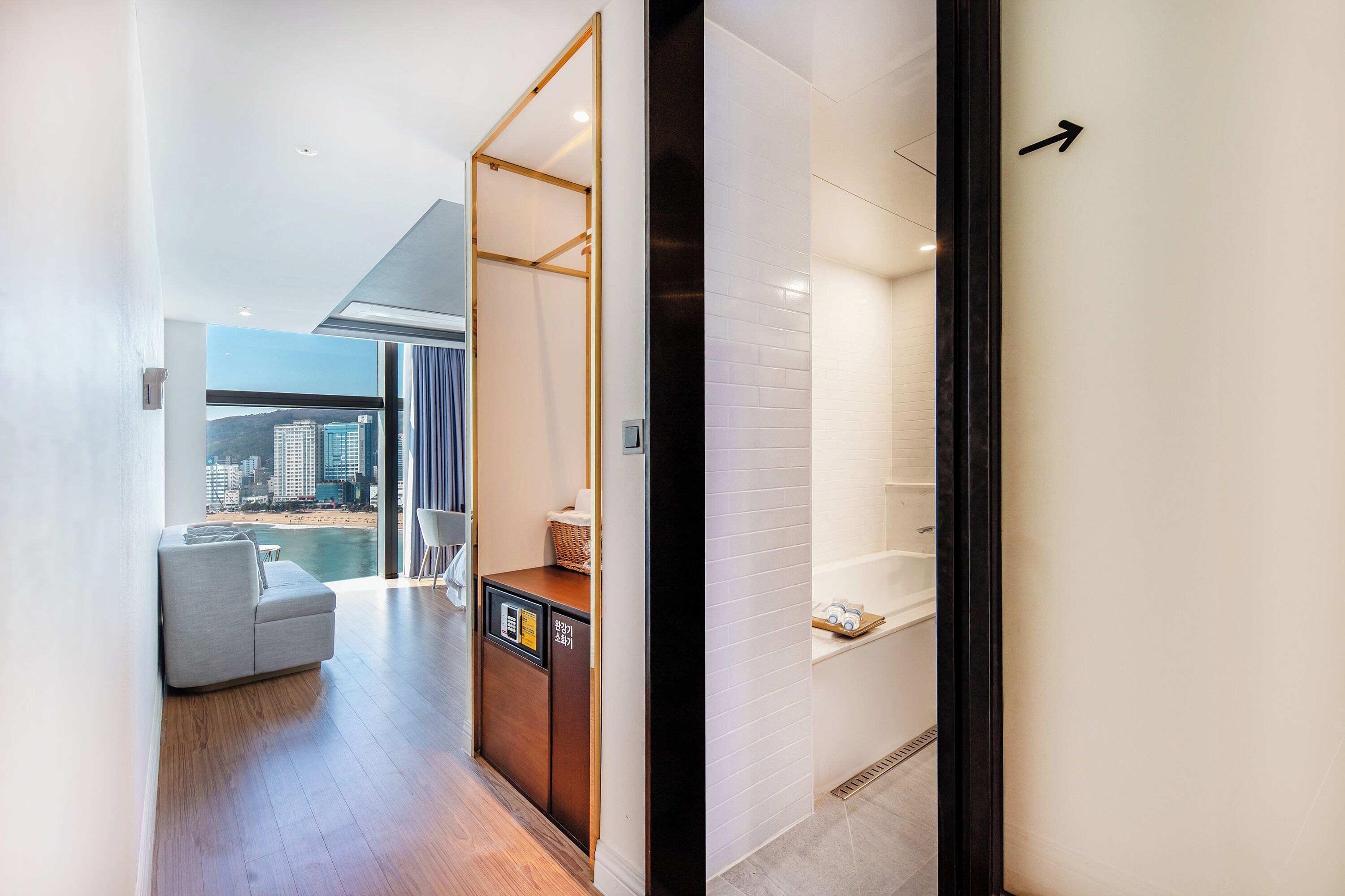 Een moderne hotelgang met een houten vloer, een grijze bank, een klein houten kastje en een stadsgezicht door grote ramen.