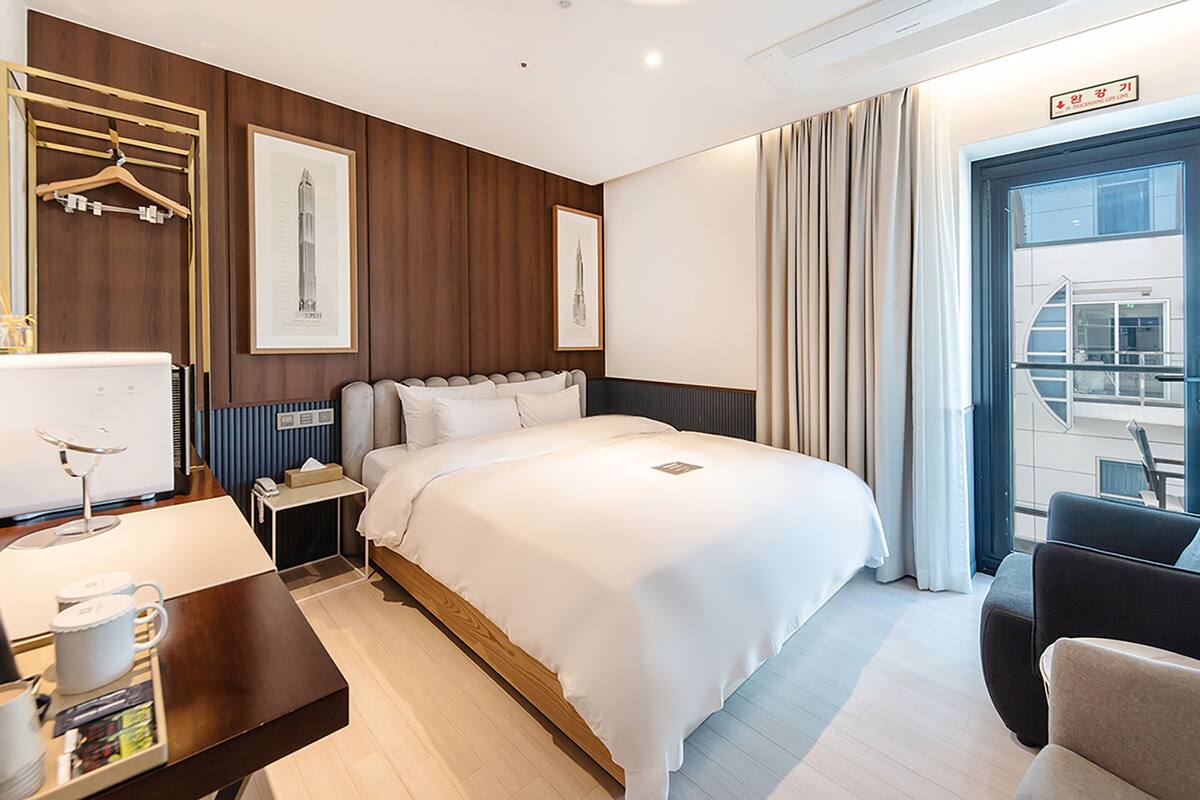 A hotel room with a large bed, bedside tables, a desk with a lamp, a chair, and a view of the city through the window.