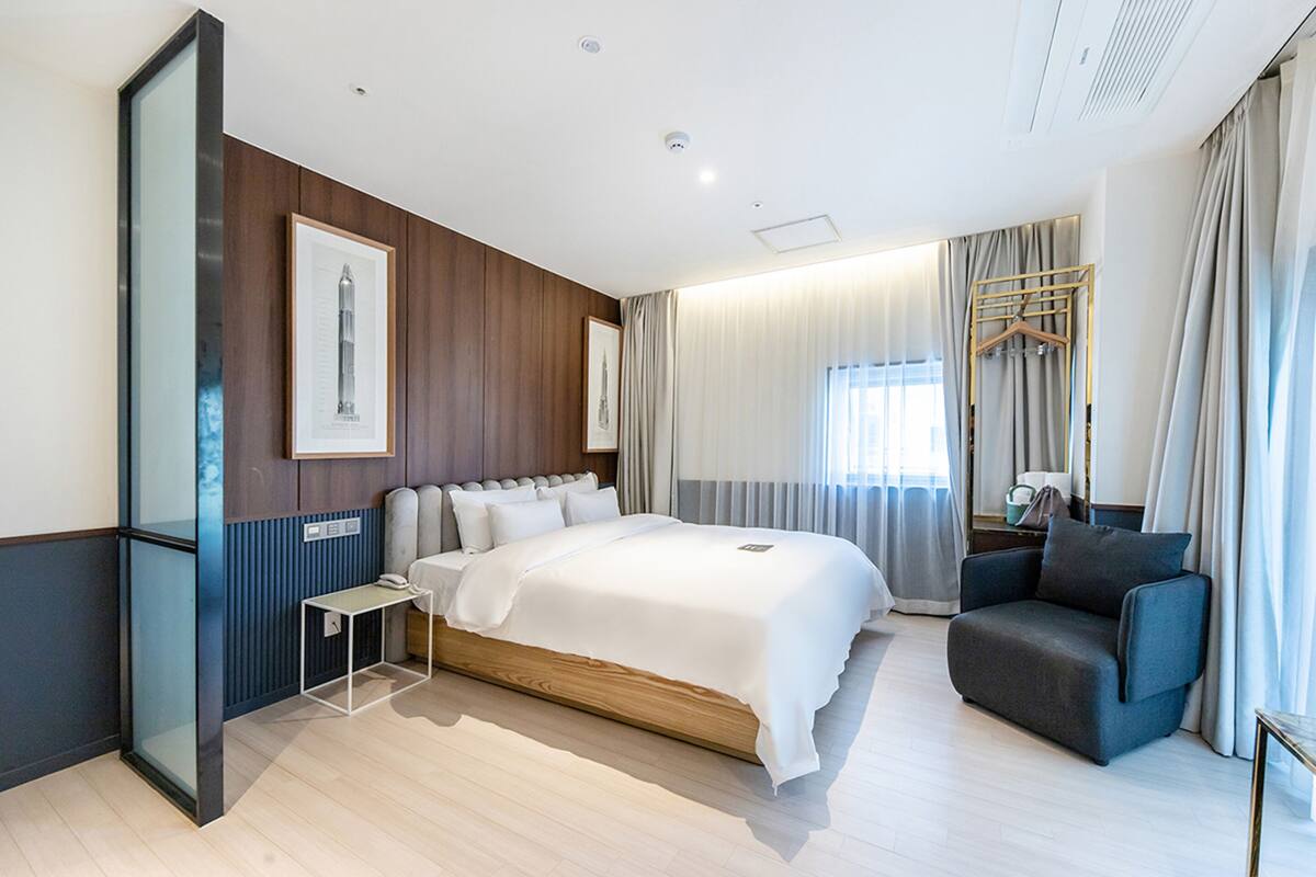 A modern hotel room with a large bed, a seating area, a small table, and a wardrobe.