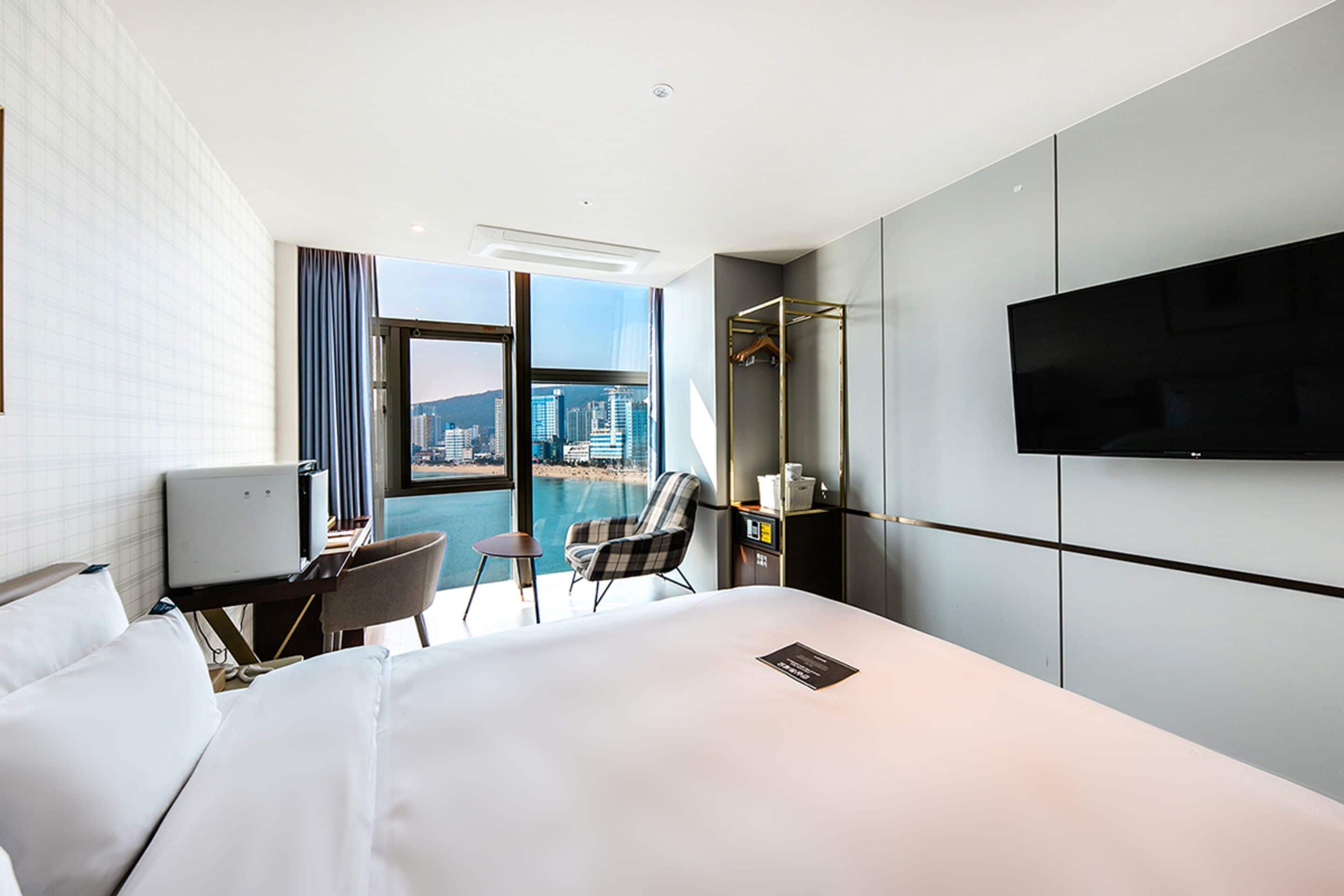 A hotel room with a large bed, a desk with a chair, a television, and a view of a cityscape through a large window.