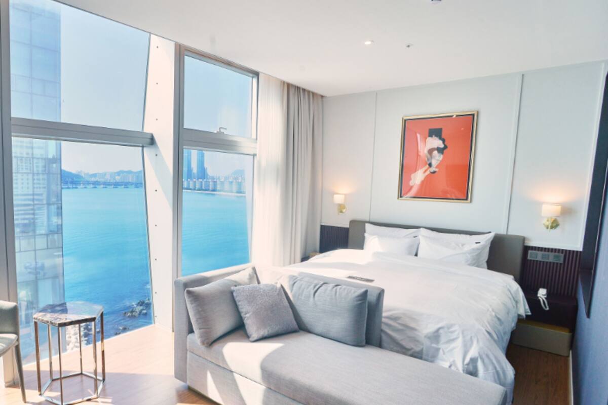 A modern hotel room with a large bed, a sofa, and a view of a cityscape through floor-to-ceiling windows.