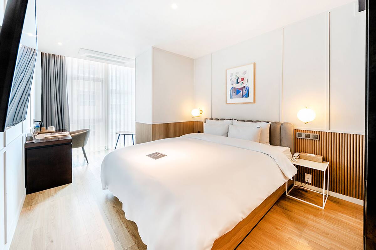 A modern hotel room with a large bed, bedside table, and a desk with a chair.