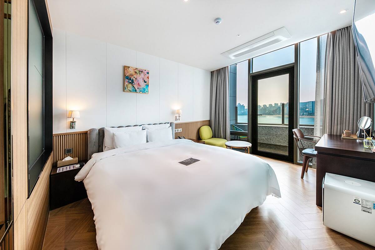 A modern hotel room with a large bed, a desk, a chair, and a view of the city through the window.