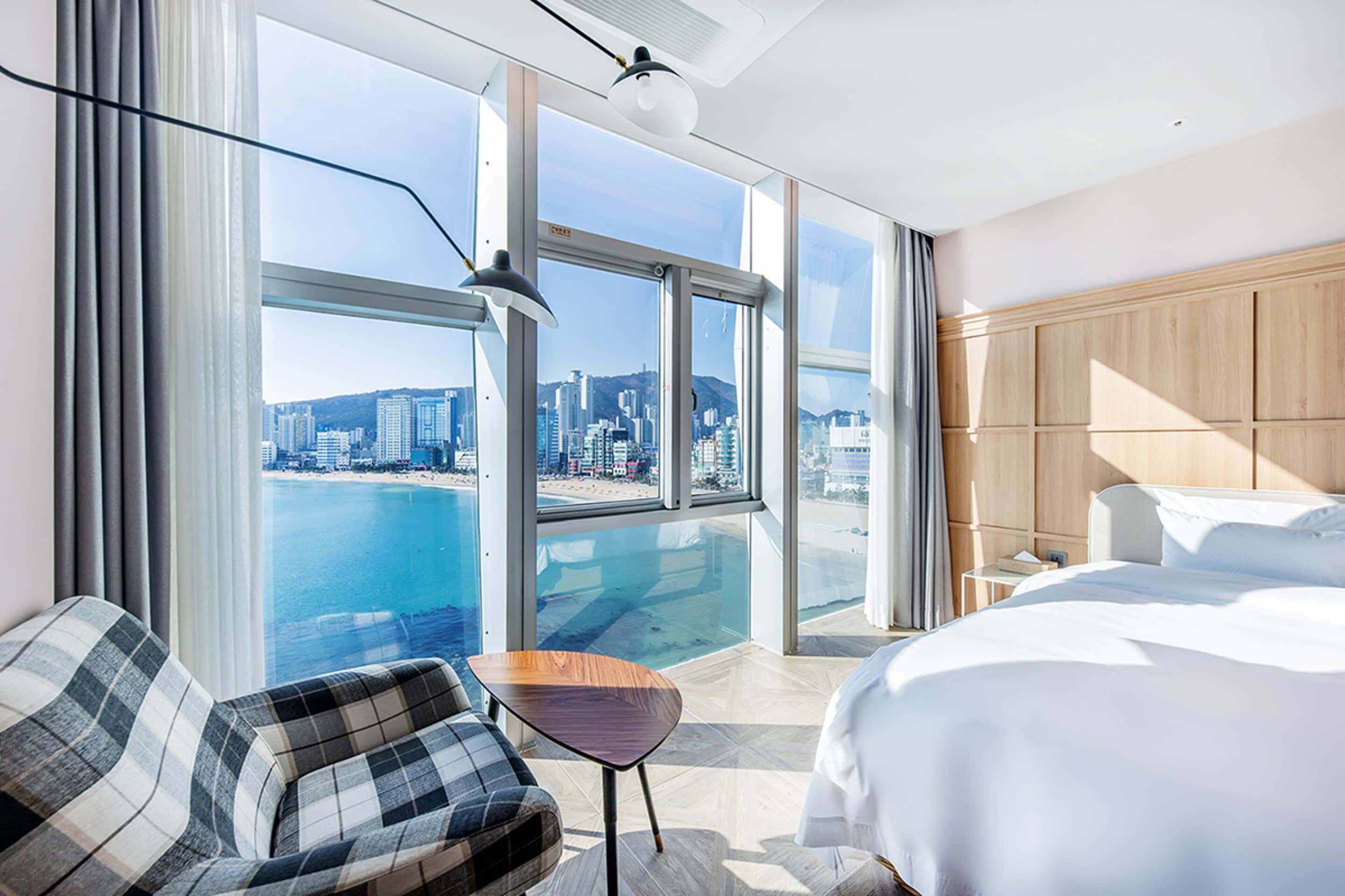 A hotel room with a large window offering a city view, a bed, a chair, and a small table.