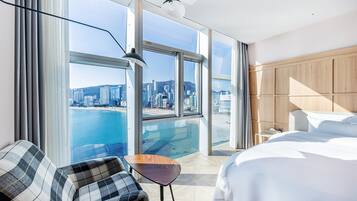 A hotel room with a large window offering a city view, a bed, a chair, and a small table.