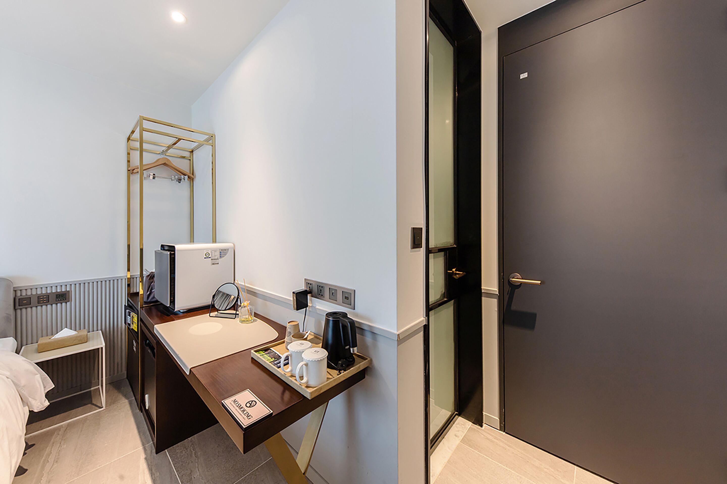 A modern hotel room with a wooden desk, a small bed, a radiator, a dark door, and a glass partition.