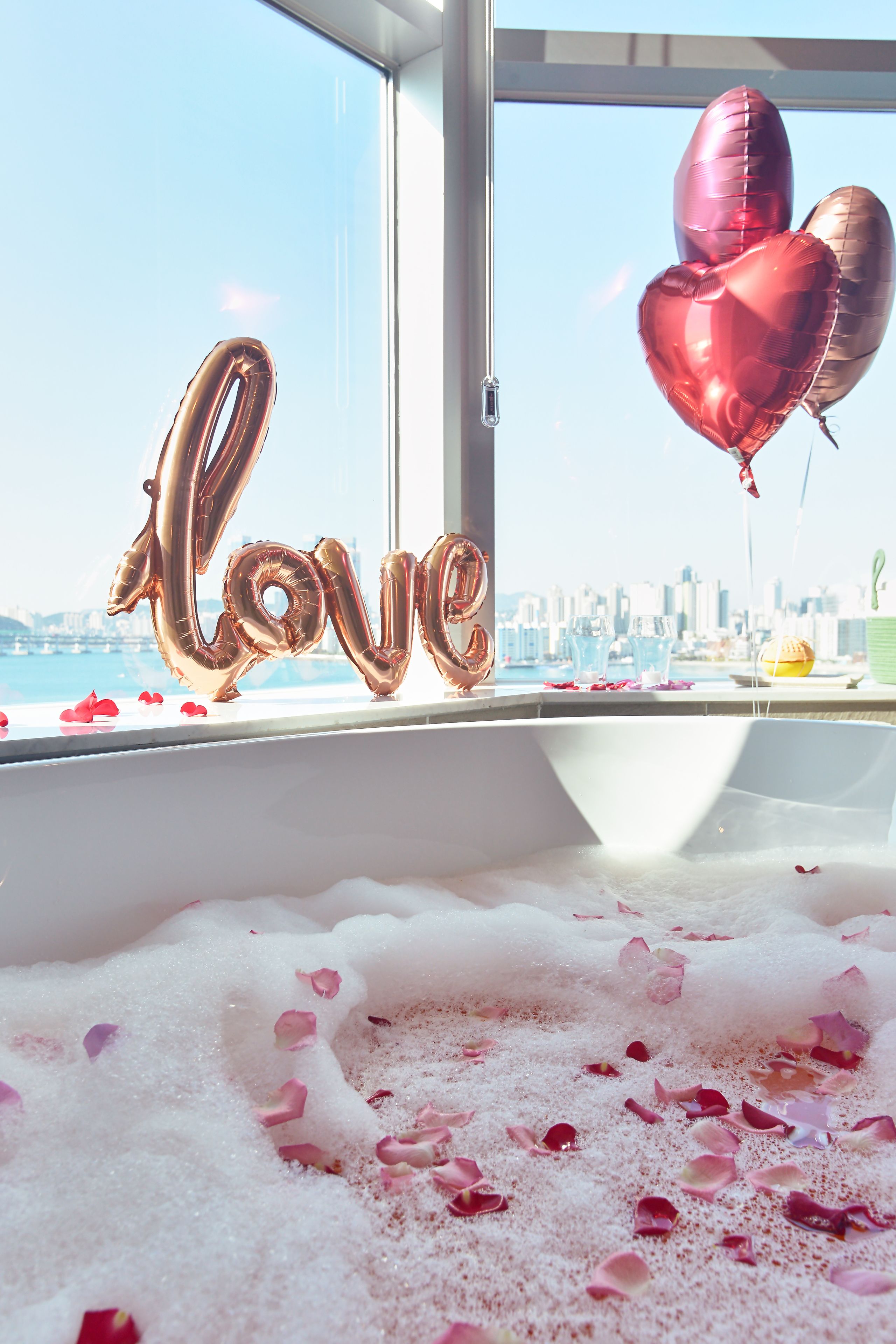 Eine Badewanne mit Rosenblüten und herzförmigen Ballons, die das Wort 'love' bilden, mit Blick auf eine Stadtlandschaft im Hintergrund.