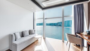 Un soggiorno moderno con un divano, un tavolino rotondo e una poltrona. Un'ampia finestra offre vista su un corpo d'acqua e sullo skyline della città.