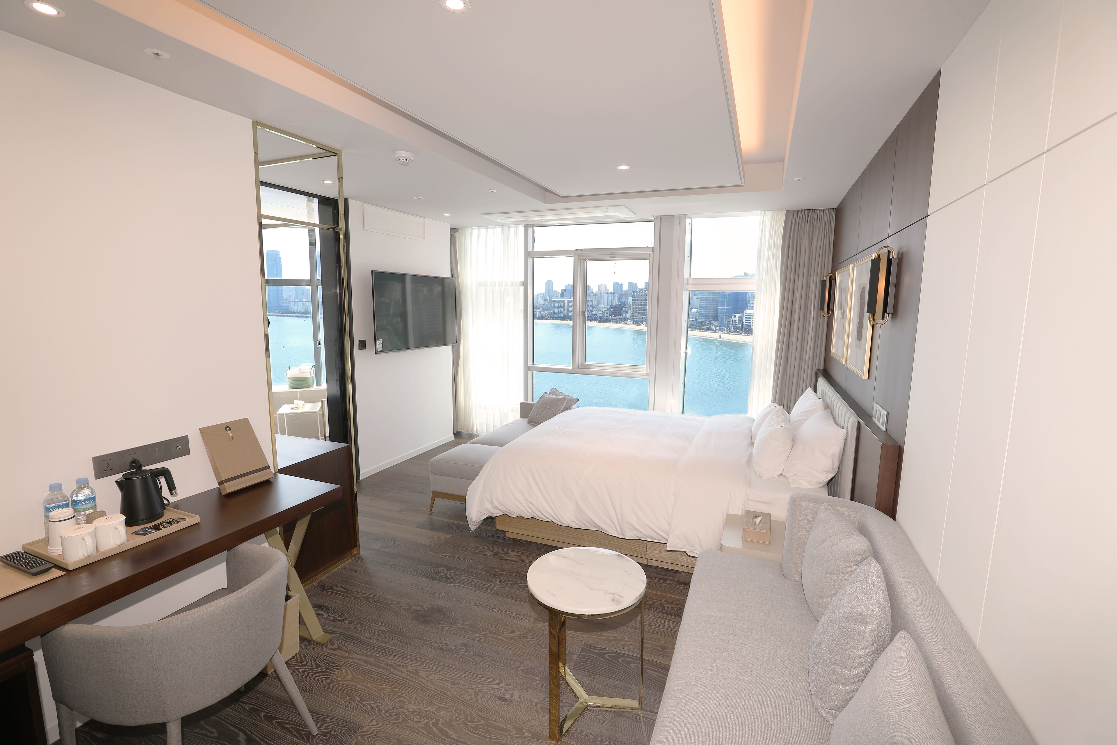 Ett modernt hotellrum med en stor säng, ett skrivbord med en stol, ett litet runt bord och utsikt över staden och vattnet.