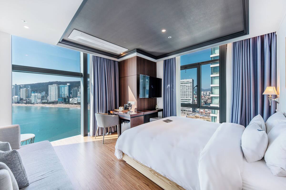 A modern hotel room with a large bed, a desk, and a TV. It has a city view through large windows.