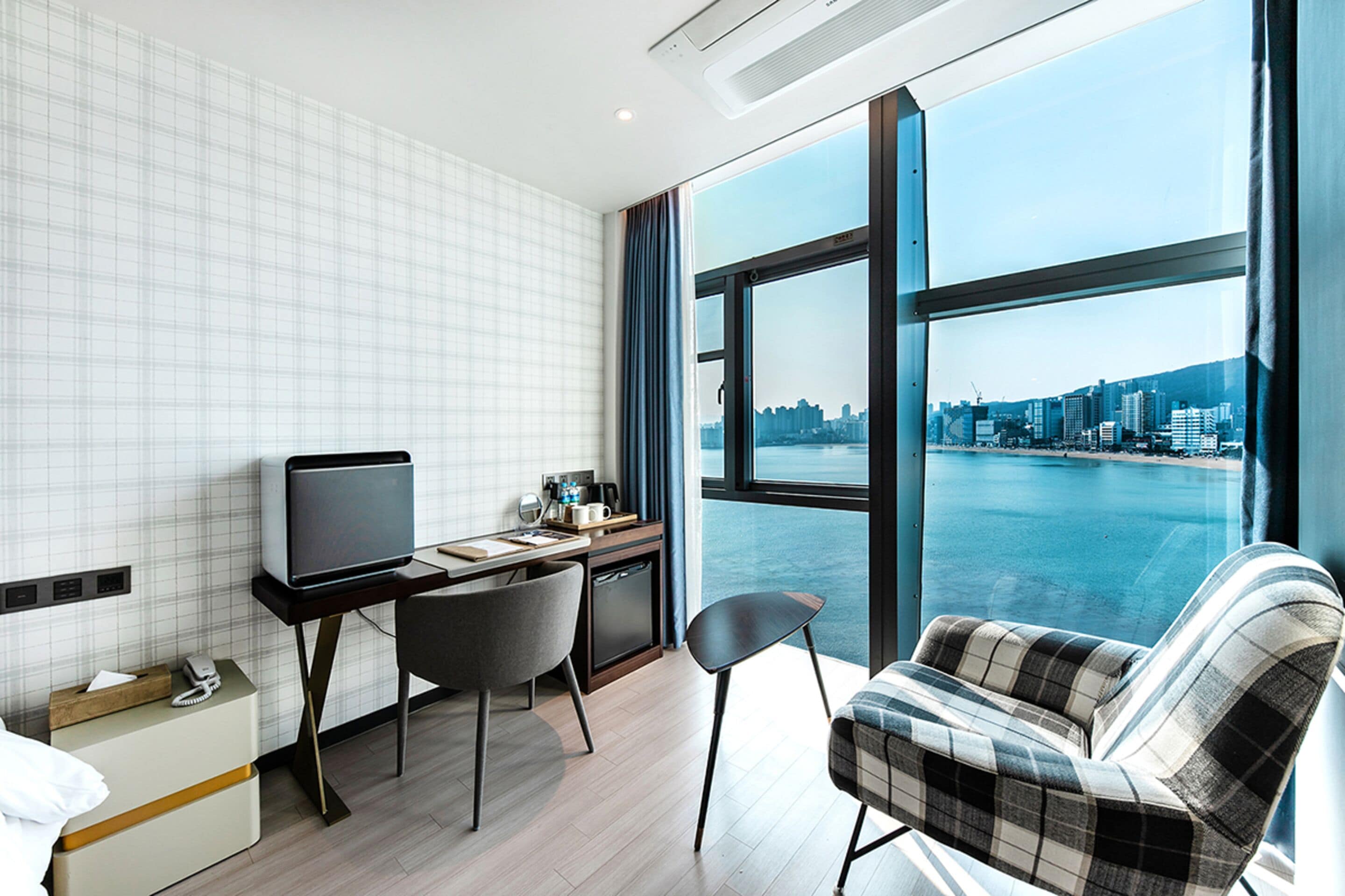 A modern hotel room with a city view, a desk with a computer, a chair, and a bed.