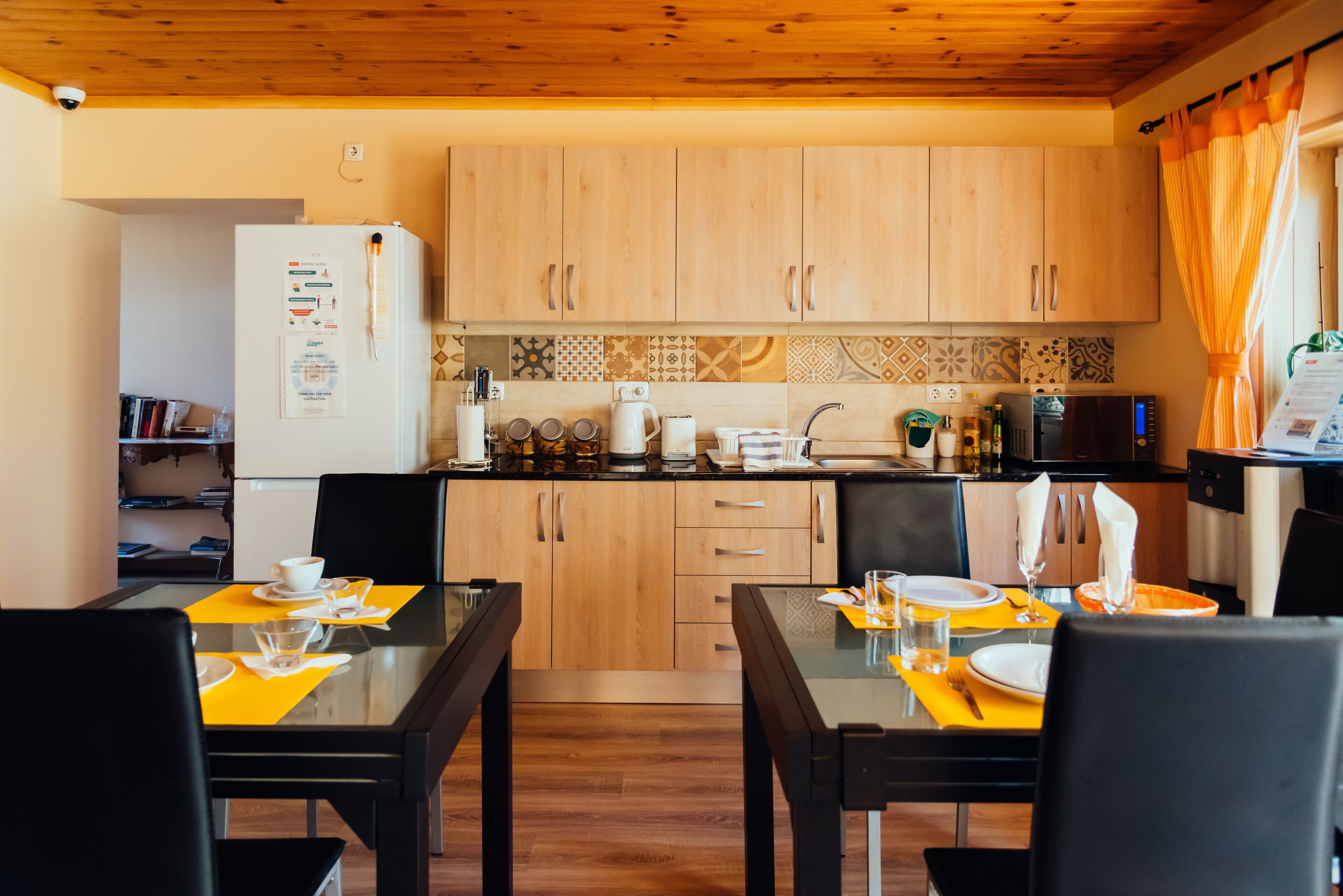 Een functionele keuken met houten kastjes, een koelkast en een magnetron. Twee eettafels met stoelen en een klein plankje met boeken en decoratieve objecten.