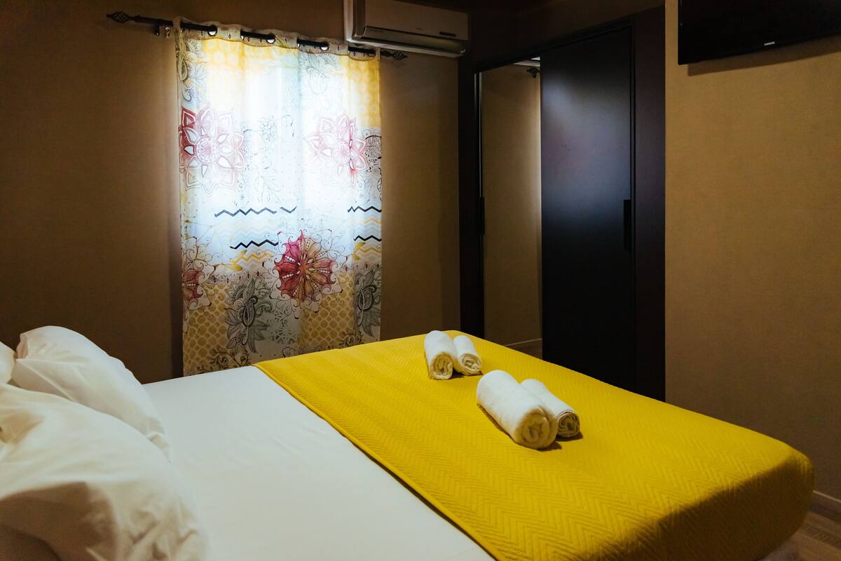 Una habitación de hotel con una cama, una colcha amarilla y dos toallas enrolladas sobre la cama.