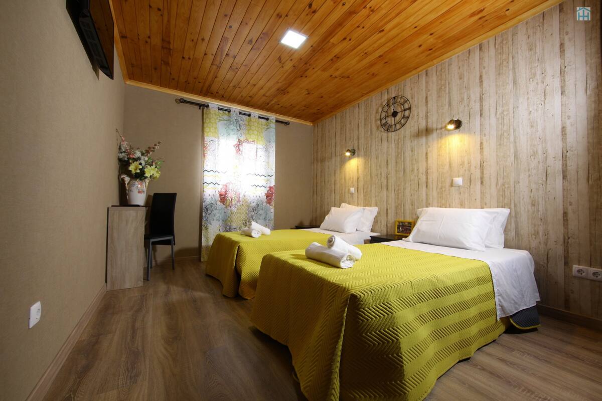 Una habitación con dos camas, piso y techo de madera, y una ventana con cortina.