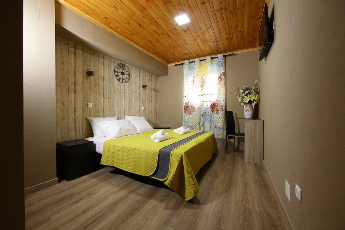Habitación de hotel con techo de madera, cama con colcha amarilla, un reloj en la pared y ventana con cortina floral.