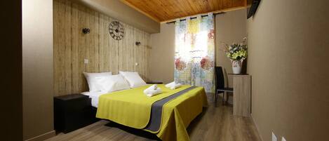 A hotel room with a wooden ceiling, a bed with a yellow bedspread, a clock on the wall, and a window with a floral curtain.
