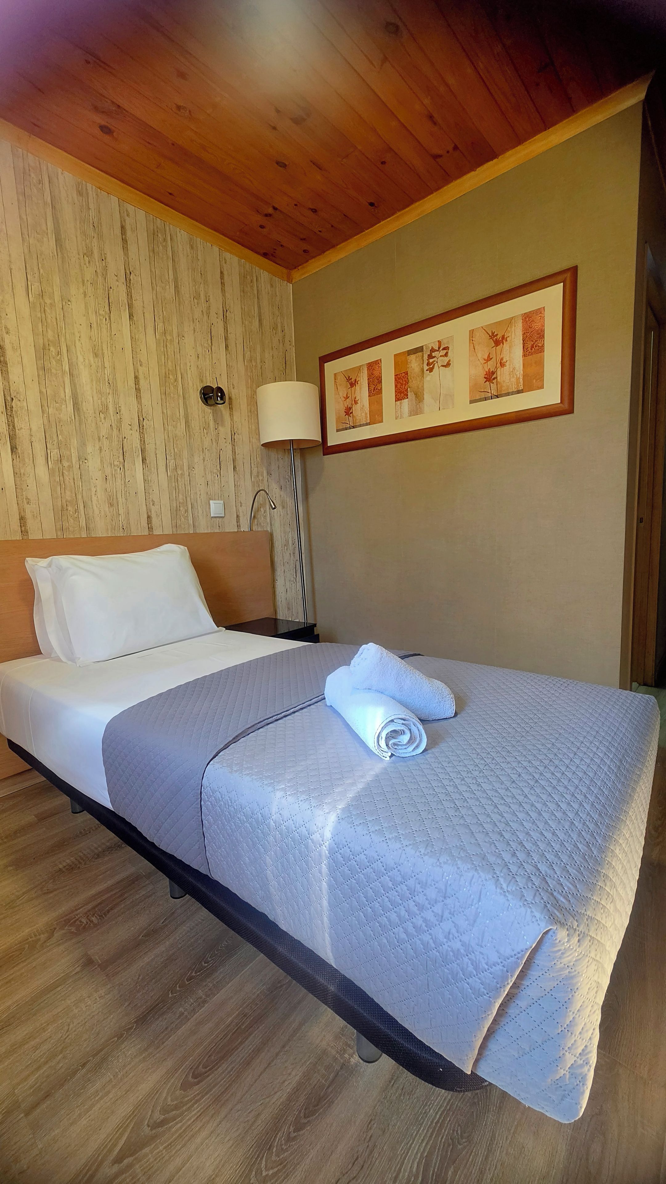 Un lit simple avec une couverture matelassée, une tête de lit en bois, une lampe fixée au mur et une œuvre d’art florale encadrée.