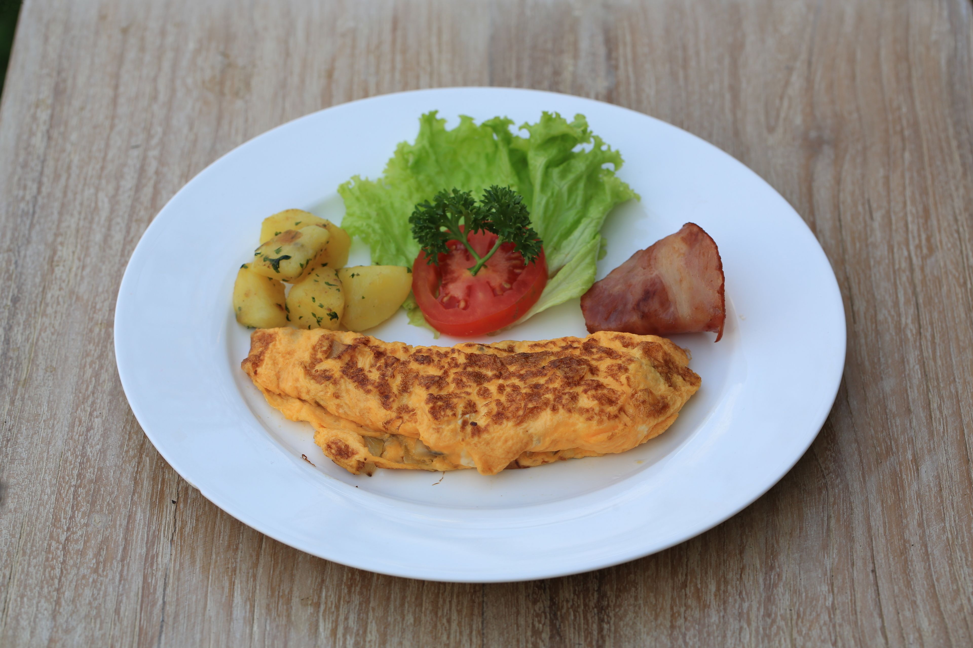 Une assiette contenant une omelette, des pommes de terre, de la laitue, une tomate et une tranche de jambon, le tout posé sur une table en bois.