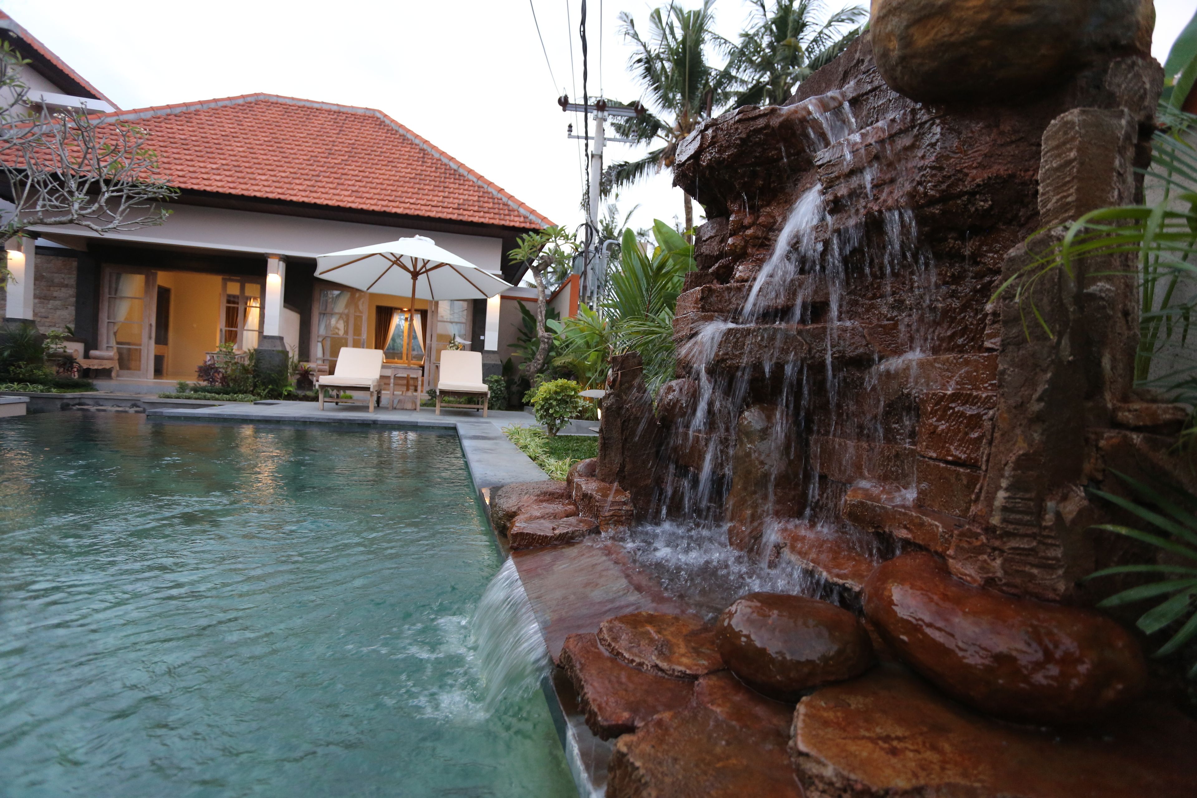 A swimming pool with a waterfall feature, surrounded by a patio area with outdoor furniture and a house in the background.