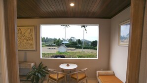 A room with a large window, a small table with chairs, a bench, and a potted plant. - Uma Dhari Villa (Sukawati)
