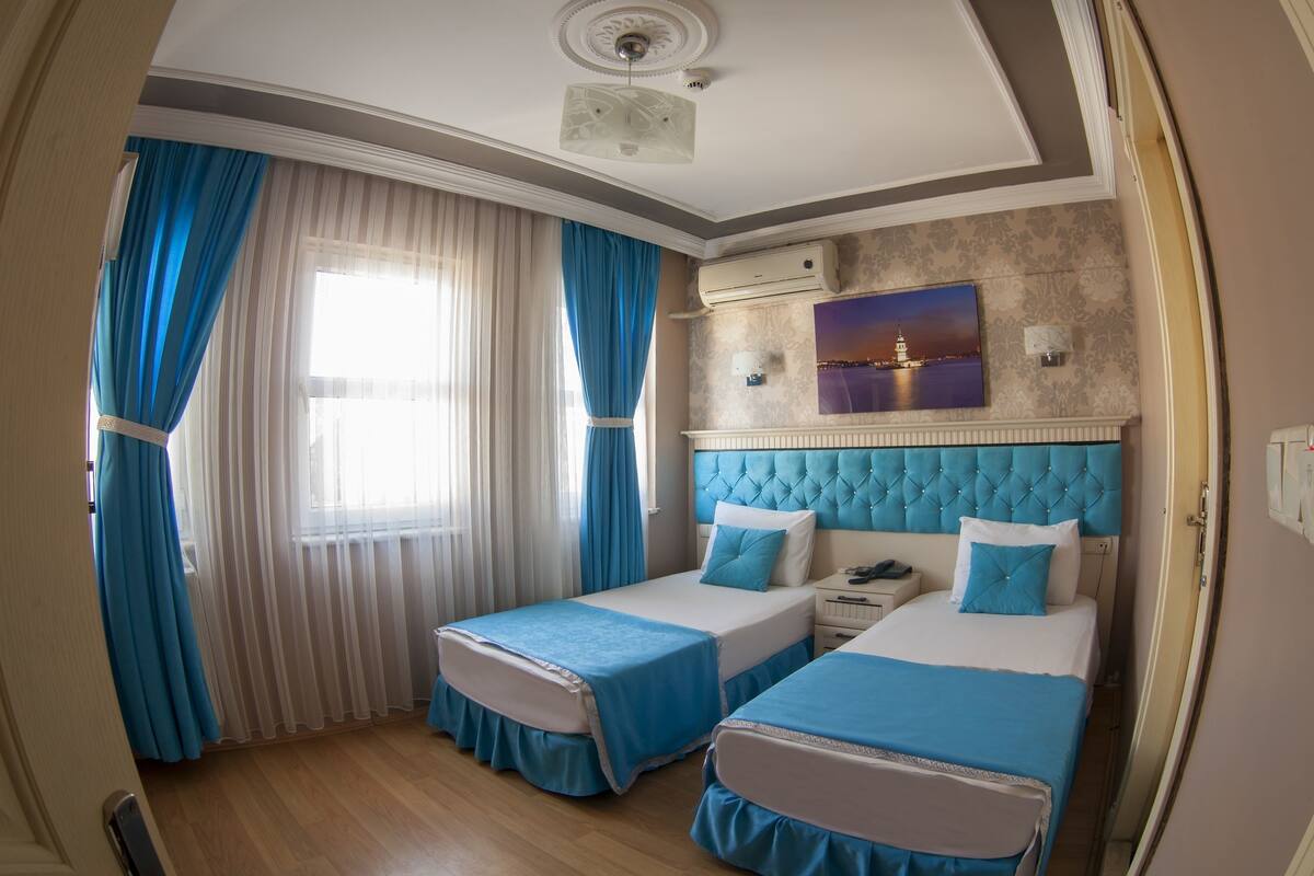 A hotel room with two beds, a large window with curtains, a painting on the wall, and an air conditioning unit.