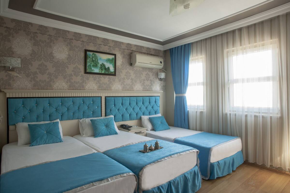 A hotel room with two beds, a large window with sheer curtains, a decorative wall, and a mounted air conditioner.