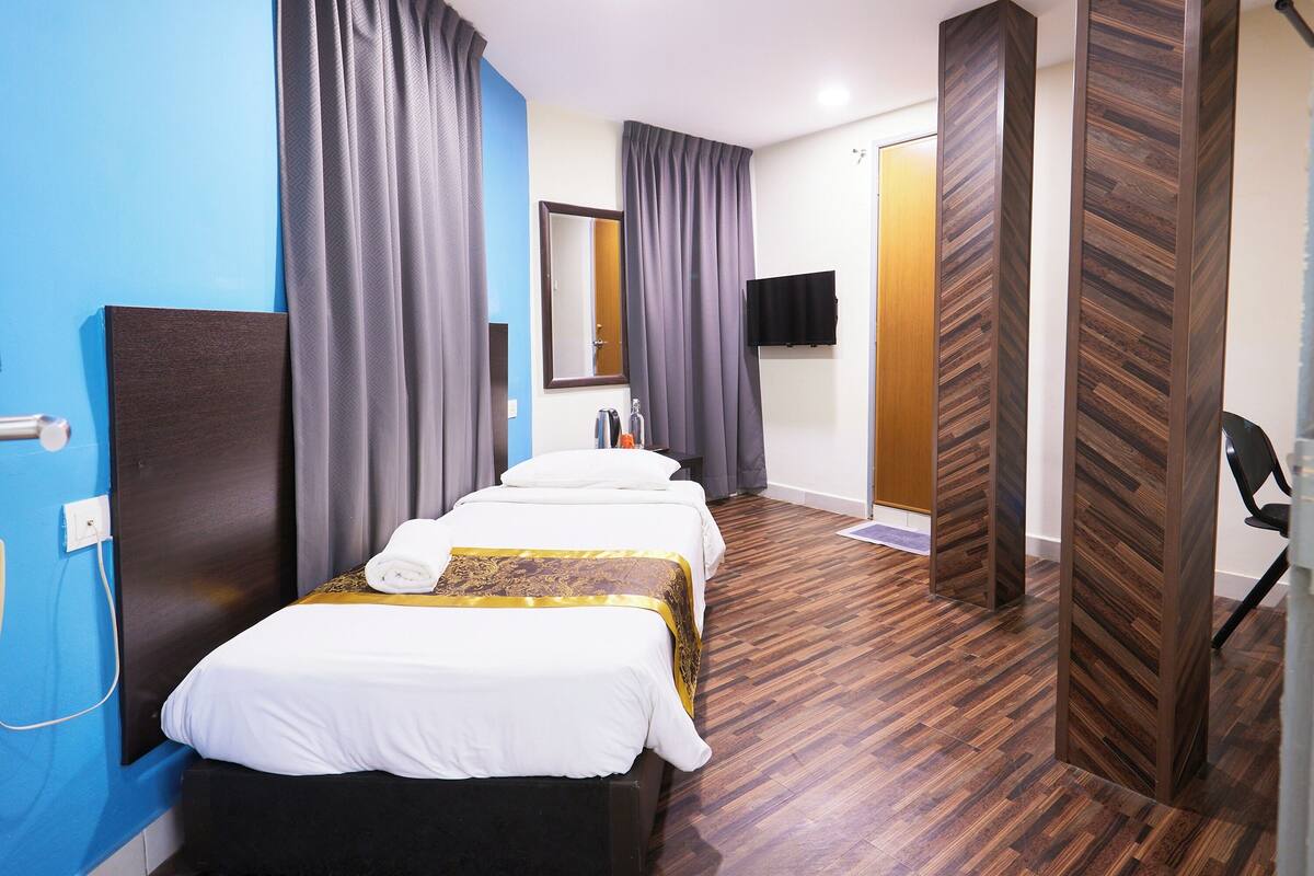 A hotel room with a blue accent wall, a wooden floor, a bed with white bedding and a gold accent, a flat-screen TV, and a door with a wooden panel design.