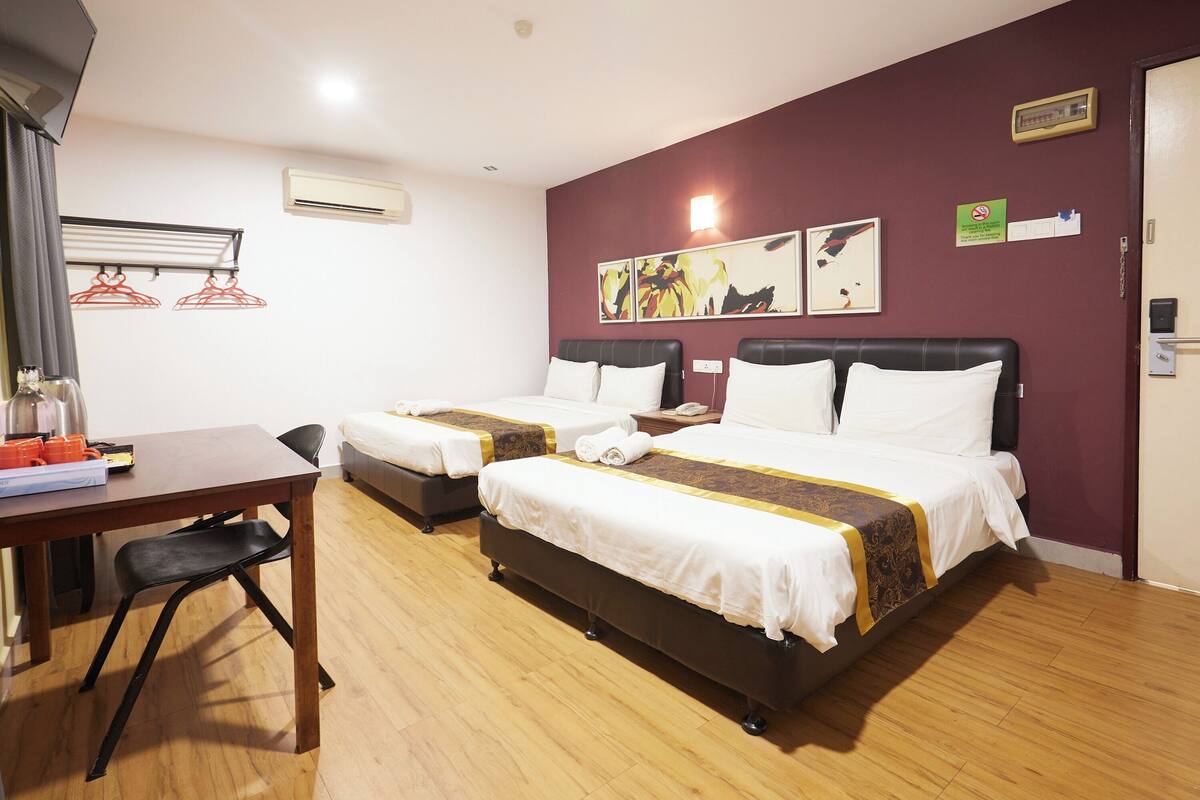 A hotel room with two beds, a desk, and a chair. There are wall-mounted air conditioning units and artwork on the walls.