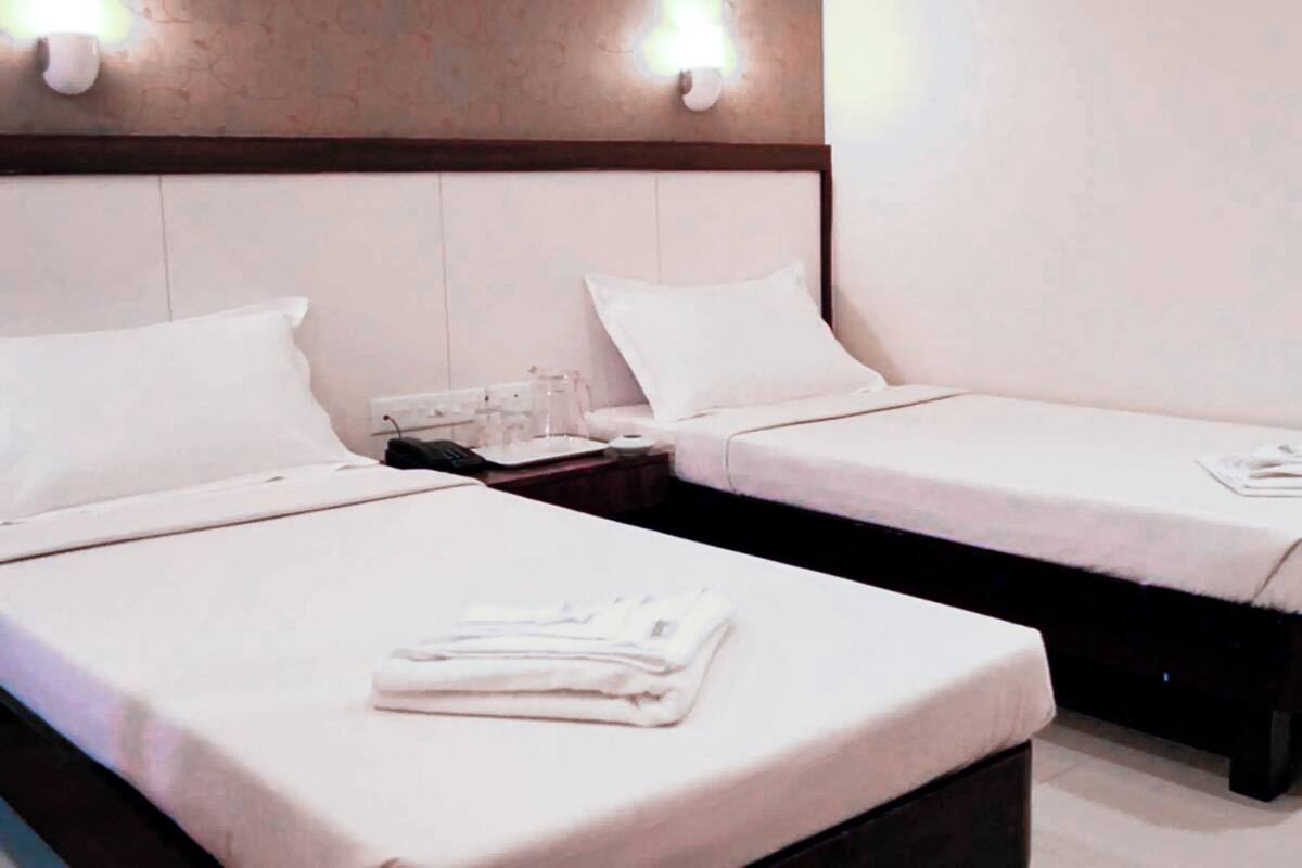 A hotel room with two single beds, each with a white bedspread and pillow, a bedside table with a lamp, and a wall-mounted light fixture.