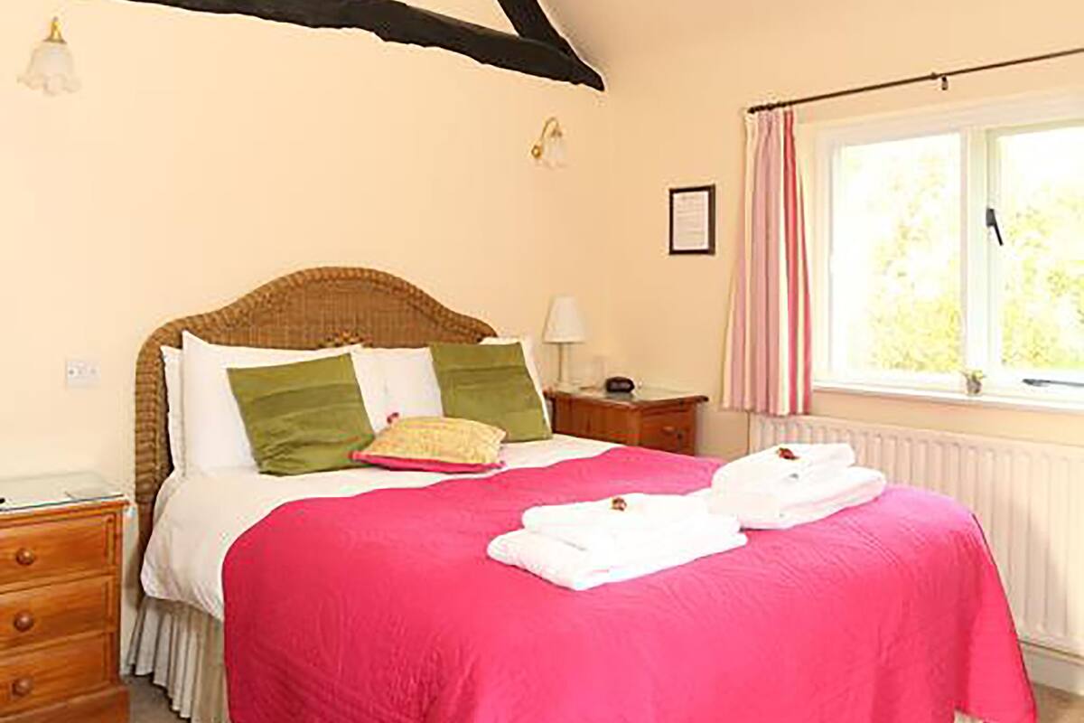 A bedroom with a bed, a nightstand, a window with curtains, and a wooden ceiling beam.