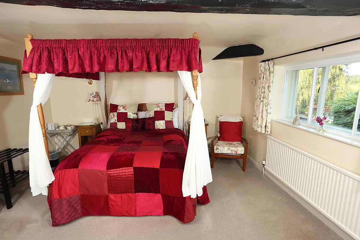 A four-poster canopy bed with a red quilt, a wooden chair, a nightstand, and a window with curtains.
