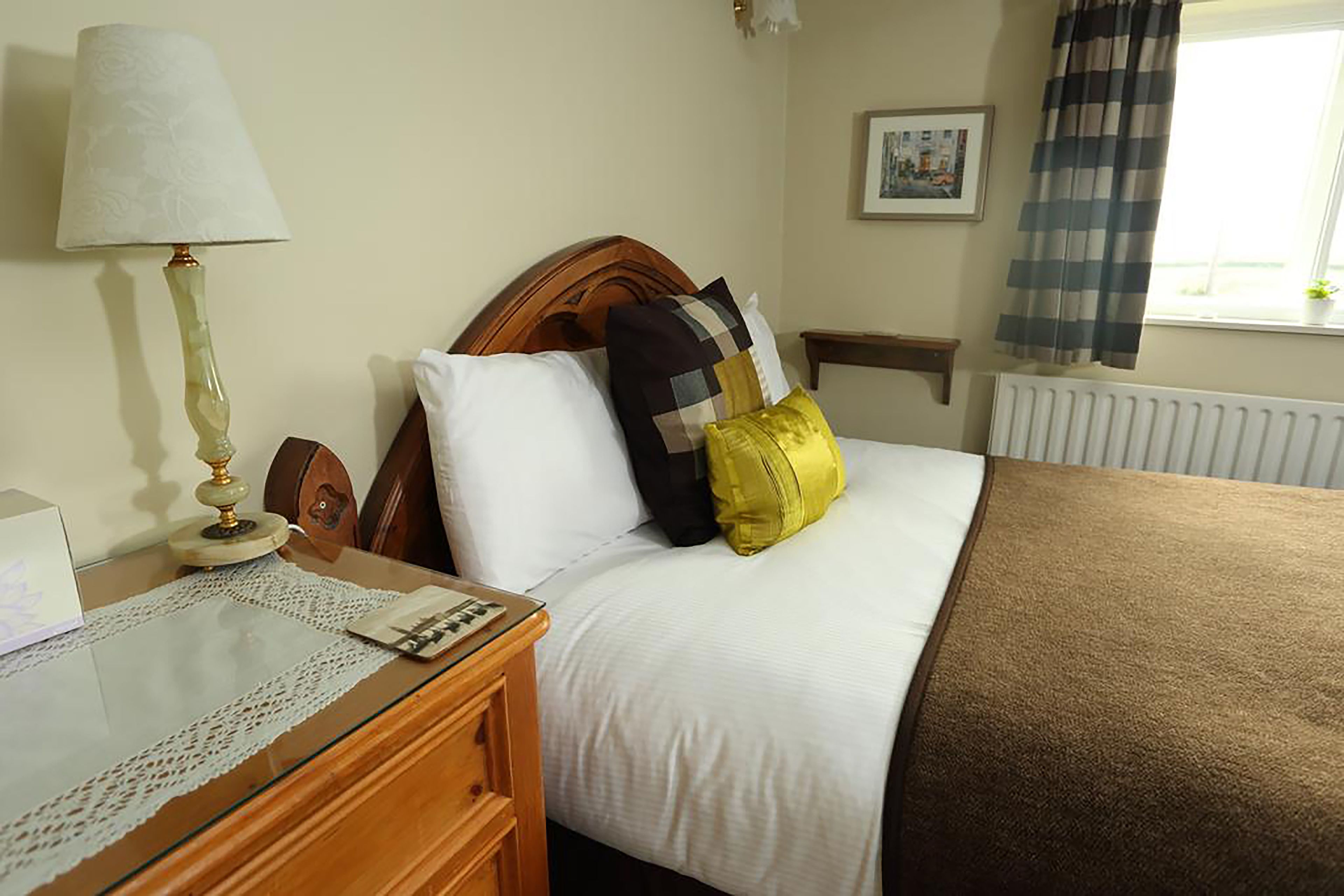A neatly made bed with a wooden headboard, a bedside table with a lamp, and a window with curtains.